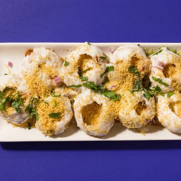 A white rectangular plate holds several Indian chaat dishes, garnished with yogurt, cilantro, and crispy noodles.