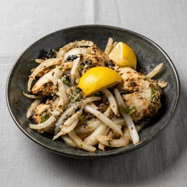 Grilled chicken pieces with sautéed onions, black olives, and lemon wedges in a dark bowl.