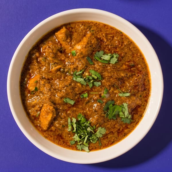 A white bowl filled with chicken curry, garnished with fresh cilantro, on a purple background.