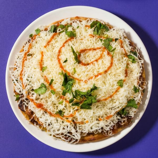 A white plate holds a savory pancake topped with shredded cheese, cilantro, and a swirl of red sauce.